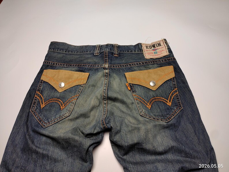 May include: Blue denim jeans with tan suede-like pocket details and orange stitching. The back pockets feature a distinctive design with a silver button closure. The Edwin brand label is visible at the waistband.