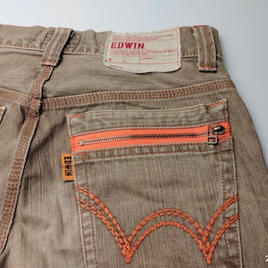 May include: Close-up of brown denim jeans with orange stitching and a zippered back pocket. The jeans feature the brand name "EDWIN" on a tag and a small orange label. The stitching and zipper provide a pop of color against the neutral denim.