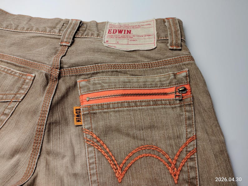 May include: Close-up of brown denim jeans with orange stitching and a zippered back pocket. The jeans feature the brand name "EDWIN" on a tag and a small orange label. The stitching and zipper provide a pop of color against the neutral denim.