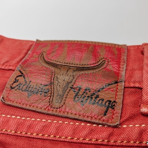 May include: Close-up of red denim fabric with a leather patch. The patch features a bull skull design and the words "Exclusive Vintage". The denim has visible stitching and a textured appearance.