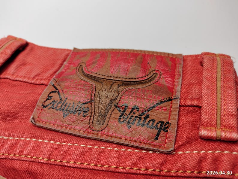 May include: Close-up of red denim fabric with a leather patch. The patch features a bull skull design and the words "Exclusive Vintage". The denim has visible stitching and a textured appearance.