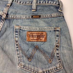 May include: Close-up of light blue denim jeans with a brown leather Wrangler patch on the back pocket. The jeans have a classic five-pocket design with visible stitching and a Wrangler label.