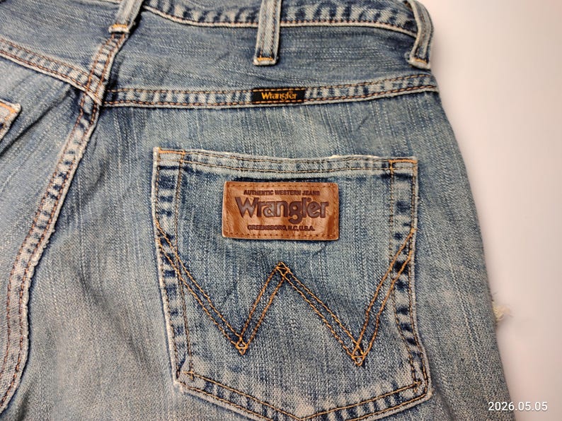 May include: Close-up of light blue denim jeans with a brown leather Wrangler patch on the back pocket. The jeans have a classic five-pocket design with visible stitching and a Wrangler label.