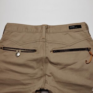 May include: Khaki-colored pants with a back pocket featuring a zipper and a decorative leather pull. The pants have a button closure and a small brand label. The image is taken from a high angle.