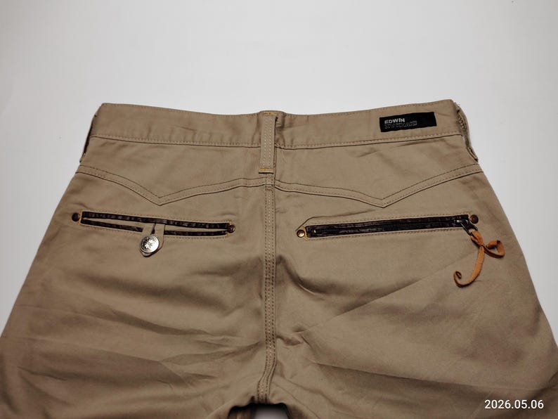 May include: Khaki-colored pants with a back pocket featuring a zipper and a decorative leather pull. The pants have a button closure and a small brand label. The image is taken from a high angle.