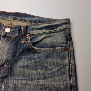 May include: Close-up of blue denim jeans with a faded wash. The jeans have a classic five-pocket design, with a small coin pocket and copper rivets. The stitching is a contrasting orange-brown color, and the jeans show signs of wear.
