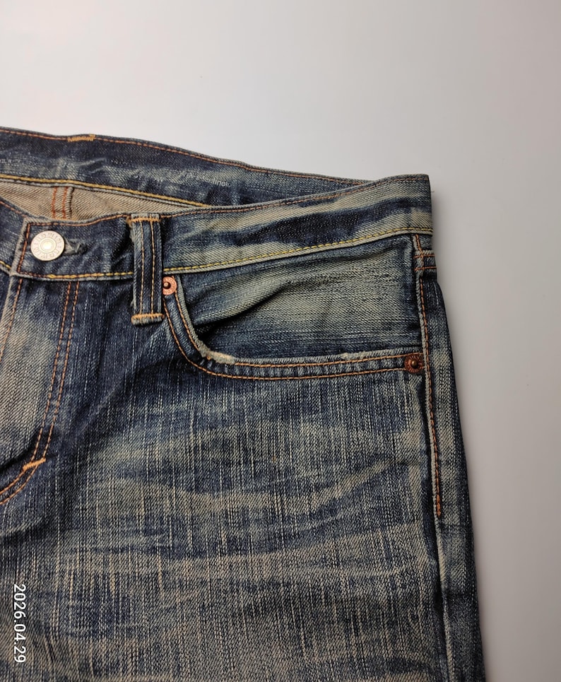 May include: Close-up of blue denim jeans with a faded wash. The jeans have a classic five-pocket design, with a small coin pocket and copper rivets. The stitching is a contrasting orange-brown color, and the jeans show signs of wear.