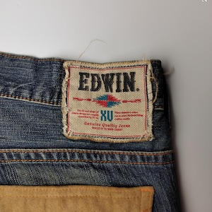 May include: Close-up of blue denim jeans with a light brown suede pocket and a decorative stitch. The jeans feature a rectangular label with the brand name "EDWIN" and the text "Genuine Quality Jeans".