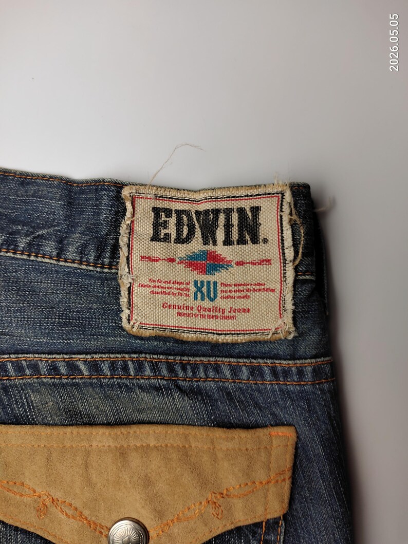 May include: Close-up of blue denim jeans with a light brown suede pocket and a decorative stitch. The jeans feature a rectangular label with the brand name "EDWIN" and the text "Genuine Quality Jeans".