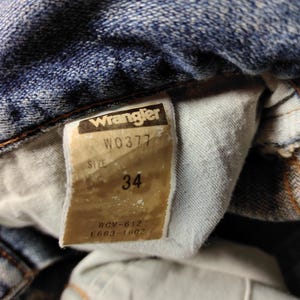 Peut inclure: Gros plan sur un jean en denim bleu avec une &eacute;tiquette Wrangler. L'&eacute;tiquette indique la taille 34. Le jean pr&eacute;sente des signes d'usure, avec des coutures et une d&eacute;coloration visibles.