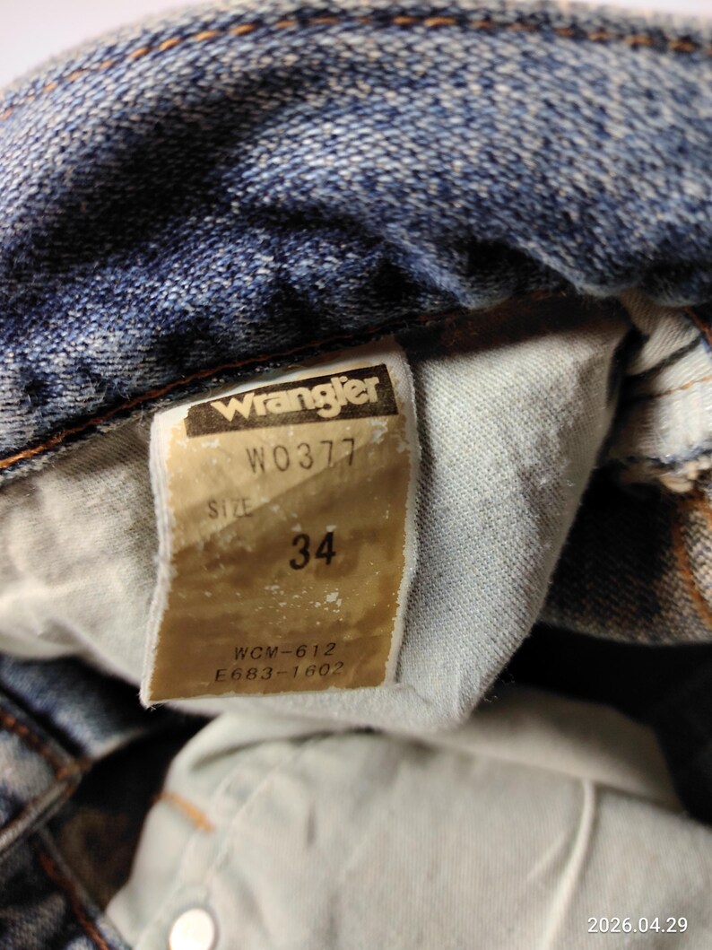 Peut inclure: Gros plan sur un jean en denim bleu avec une &eacute;tiquette Wrangler. L'&eacute;tiquette indique la taille 34. Le jean pr&eacute;sente des signes d'usure, avec des coutures et une d&eacute;coloration visibles.