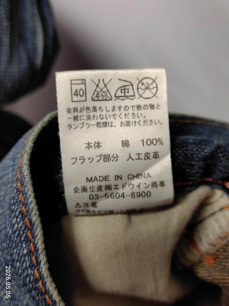 May include: Close-up of a tag on denim jeans. The tag includes washing instructions, fabric composition (100% cotton), and manufacturing details. The text is in Japanese and English.