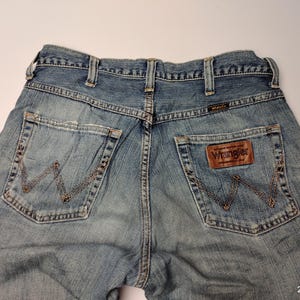 May include: Light blue denim Wrangler jeans. The jeans have a classic five-pocket design with the Wrangler logo on the back pocket and a leather patch. The stitching is a contrasting brown color.