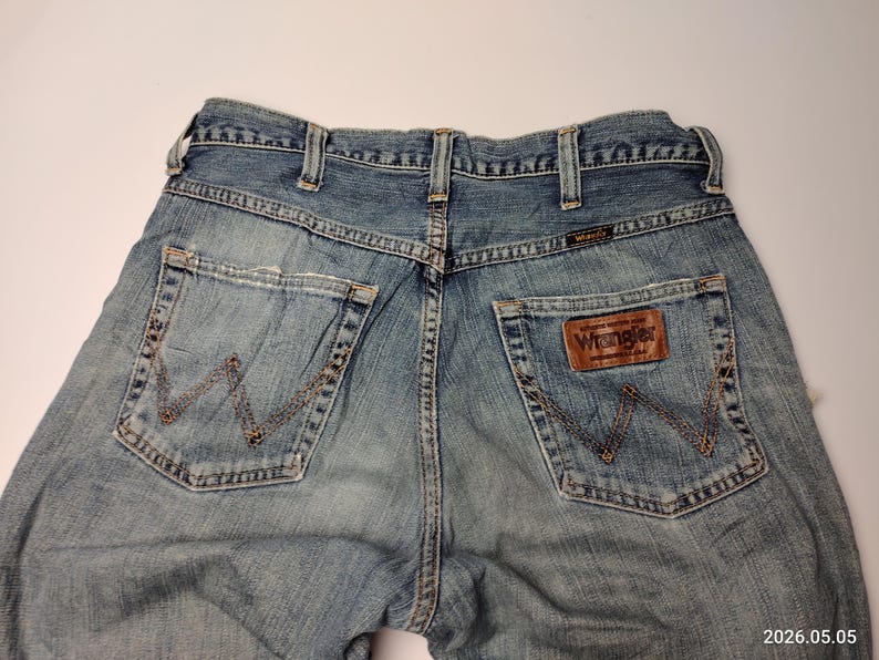 May include: Light blue denim Wrangler jeans. The jeans have a classic five-pocket design with the Wrangler logo on the back pocket and a leather patch. The stitching is a contrasting brown color.