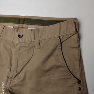 May include: Close-up of khaki-colored pants. The pants feature a button closure, belt loops, and a pocket with rivets. The stitching is visible in a contrasting color. The pants are made of a durable fabric, suitable for casual wear.