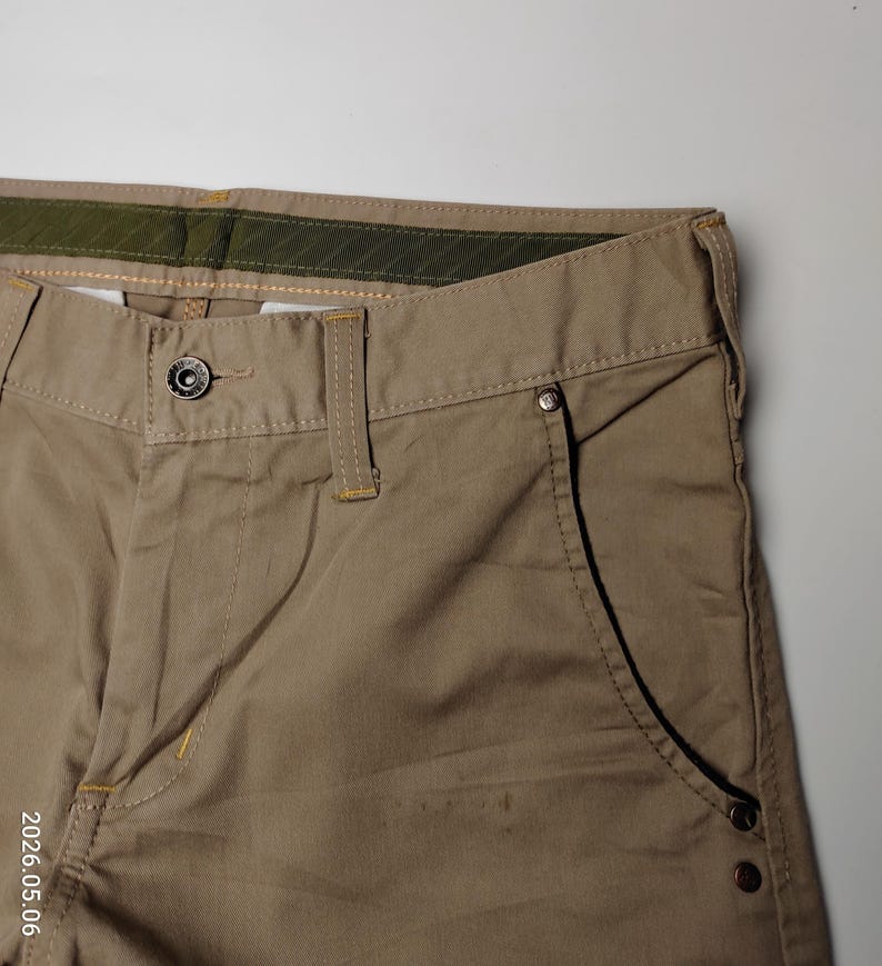 May include: Close-up of khaki-colored pants. The pants feature a button closure, belt loops, and a pocket with rivets. The stitching is visible in a contrasting color. The pants are made of a durable fabric, suitable for casual wear.