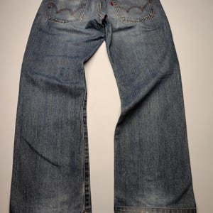May include: Faded blue denim jeans with classic five-pocket styling. Features include a red tab on the back pocket and a brown leather patch at the waistband. The jeans have a relaxed fit and show signs of wear.