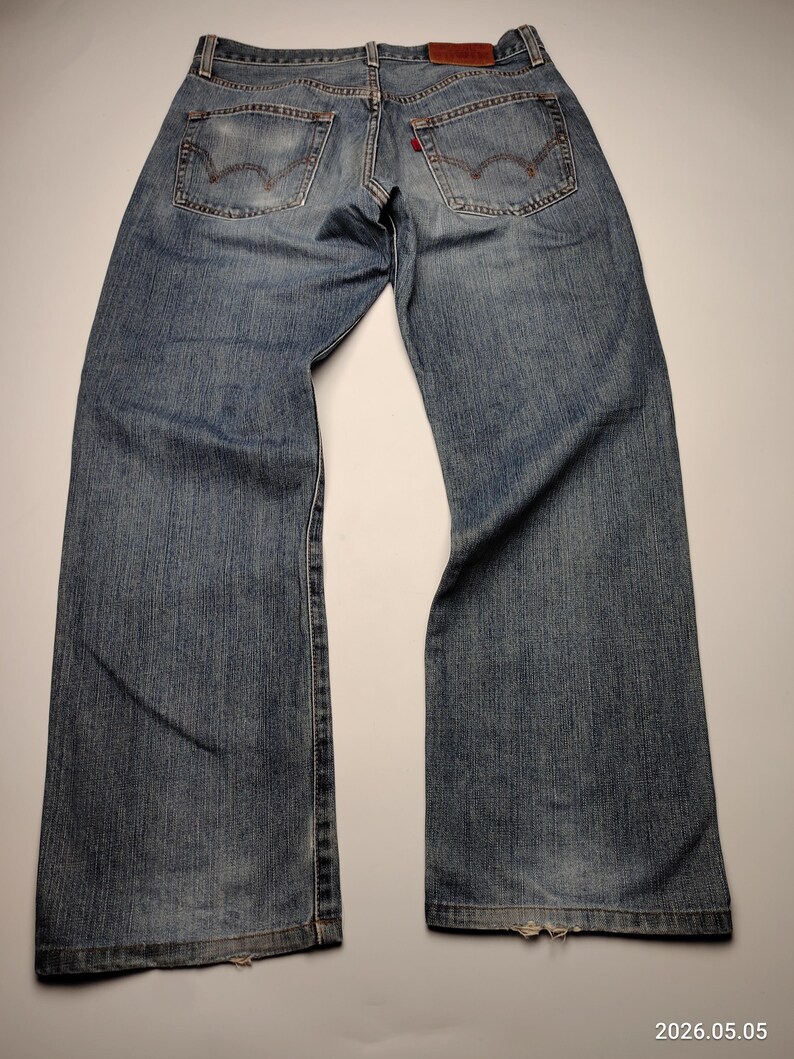 May include: Faded blue denim jeans with classic five-pocket styling. Features include a red tab on the back pocket and a brown leather patch at the waistband. The jeans have a relaxed fit and show signs of wear.