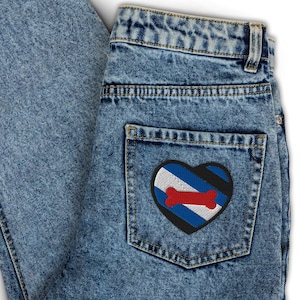 May include: Light blue denim jeans with a heart-shaped patch on the back pocket. The patch features a red bone design over blue and white stripes, framed by a black border.