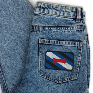 May include: Light blue denim jeans with a patch on the back pocket. The patch features a design with blue, white, black, and a red bone shape. The jeans have a classic five-pocket design.