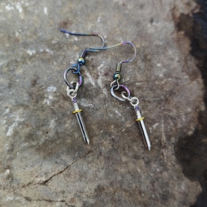 May include: A pair of handmade earrings featuring small, silver-colored sword charms. The earrings have a hook design with a metallic, iridescent finish. The sword charms have a gold-colored hilt and a silver blade. The earrings are displayed on a textured, gray surface.