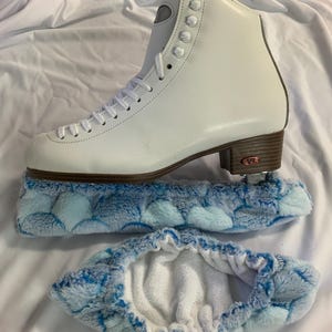 Puede incluir: Un patín de figura blanco con cordones y un tacón marrón, apoyado sobre un protector de patín y una funda de cuchilla con estampado azul y blanco. El patín tiene un diseño clásico con ojales metálicos y un cuello acolchado.