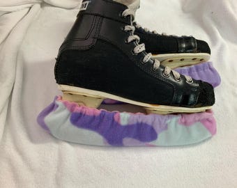 May include: Black ice skates with white laces and a black toe cap, resting on a purple and pink patterned skate guard. The skates are on a white surface.