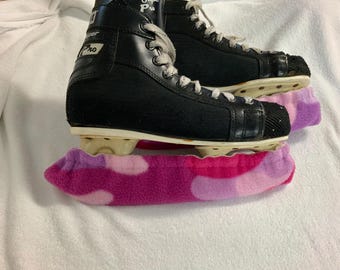 May include: A pair of black ice skates with white laces and a white sole, resting on a pink, purple, and white camouflage skate guard. The skates are on a white surface.