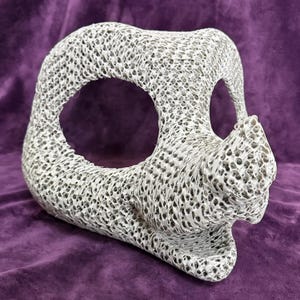 May include: A three-dimensional, off-white sculptural object featuring a complex, porous, web-like lattice structure. The form includes a large, hollow circular opening on the side and a protruding, pointed snout-like element at the front.