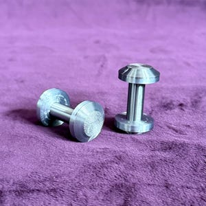 May include: Two silver-colored cufflinks with a textured, circular design on the face. The cufflinks have a cylindrical post connecting the two circular faces. One cufflink is lying on its side, while the other stands upright.