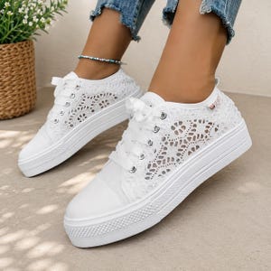 Può includere: Un paio di scarpe da ginnastica bianche con dettagli in pizzo, lacci bianchi e una spessa suola bianca. Le scarpe da ginnastica hanno un design a taglio basso. Le scarpe sono indossate da una persona che indossa jeans blu.