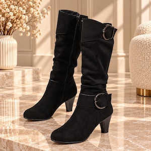 May include: A pair of black knee-high boots featuring a soft, matte finish, a rounded toe, and a mid-height block heel. Each boot includes a decorative strap across the ankle and upper calf, secured with a circular metallic buckle and a side zipper.