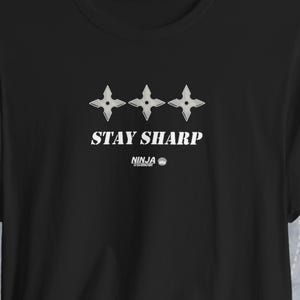 Puede incluir: Una camiseta negra de manga corta con un gráfico blanco de tres estrellas shuriken dispuestas horizontalmente sobre el texto STAY SHARP y el texto más pequeño NINJA SHADOW impreso en blanco en el centro del pecho.