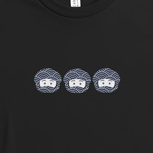 May include: Black t-shirt featuring three circular designs. Each design has a white face with eyes and a mouth, surrounded by a blue and white wave pattern. The shirt is a simple, casual style.