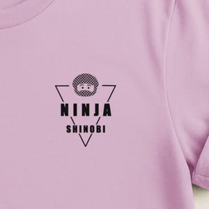 Puede incluir: Una camiseta de color lila claro con un diseño gráfico negro. El diseño incluye la palabra "NINJA" encima de "SHINOBI" en una fuente estilizada, con un círculo punteado y un icono de cara encima de la palabra "NINJA".