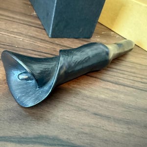 May include: A black calla lily bottle stopper with a brass stem. The stopper is made of a dark, textured material and is designed to resemble a calla lily flower. The stopper is lying on a wooden surface.