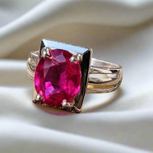 May include: A gold ring with a large, faceted, ruby red gemstone set in a square bezel. The band is a simple, polished gold.