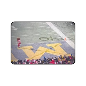Può includere: Un tappetino per mouse rettangolare con un design da campo da football e un grande logo giallo "W". Una mascotte in abito rosso e bianco è sul campo. Una folla di spettatori è visibile in primo piano.
