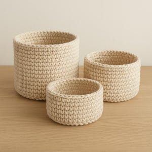 Puede incluir: Tres cestas de ganchillo color crema, de diferentes tamaños, están dispuestas sobre una superficie de madera clara. Las cestas son cilíndricas con un aspecto texturizado y hecho a mano, adecuadas para almacenamiento o decoración.