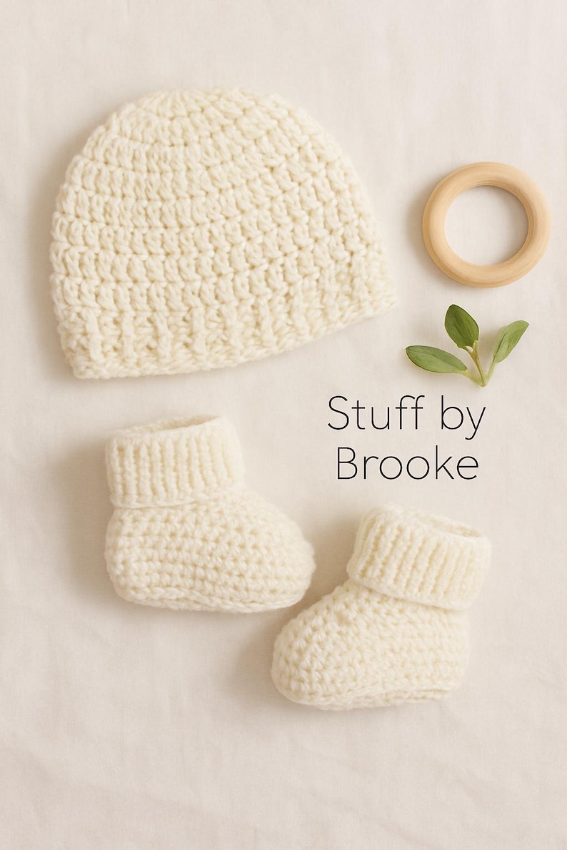 Pode incluir: Conjunto de beb&ecirc; de croch&ecirc; creme, incluindo um gorro e dois sapatinhos. Um anel de madeira e um raminho de vegeta&ccedil;&atilde;o tamb&eacute;m est&atilde;o na imagem. O texto "Stuff by Brooke" &eacute; exibido.