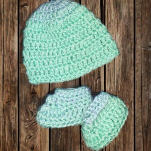 Pode incluir: Conjunto de gorro e botinhas de croch&ecirc; para beb&ecirc; em tons de verde menta e azul claro. O conjunto feito &agrave; m&atilde;o &eacute; exibido em um fundo de madeira r&uacute;stica, mostrando a textura e os detalhes do fio.