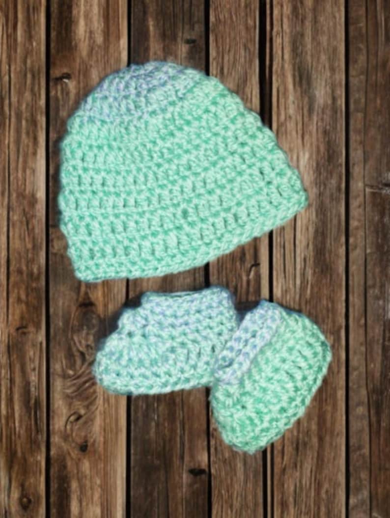 Pode incluir: Conjunto de gorro e botinhas de croch&ecirc; para beb&ecirc; em tons de verde menta e azul claro. O conjunto feito &agrave; m&atilde;o &eacute; exibido em um fundo de madeira r&uacute;stica, mostrando a textura e os detalhes do fio.