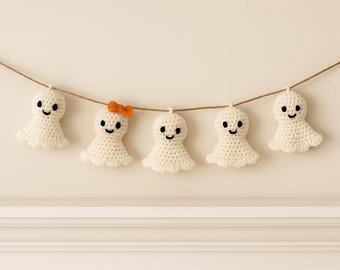 May include: A handmade Halloween garland featuring five crocheted ghost figures. Each ghost is white with a smiling face and a scalloped bottom edge. One ghost has an orange bow. The garland is strung on a brown twine.