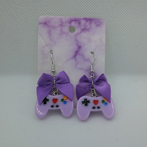 May include: A pair of lavender-colored earrings shaped like video game controllers. Each earring features a purple bow at the top and colorful buttons. The earrings hang from silver-colored fish hook ear wires.