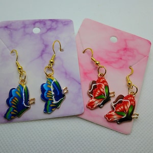 May include: Two pairs of butterfly earrings on display cards. One pair features blue and green wings, the other red and green. Both have gold-colored hooks and butterfly body details. The cards are purple and pink.