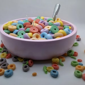 May include: A light pink bowl filled with colorful, ring-shaped cereal. A silver spoon rests in the cereal. Some cereal pieces are scattered around the bowl on a white surface. The cereal pieces are in various pastel colors.