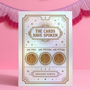 Peut inclure: Une carte blanche avec des accents dorés et le texte "THE CARDS HAVE SPOKEN." La carte comporte trois cercles à gratter avec les mentions "Le Passé", "Le Présent" et "Le Futur". La carte est ornée d'étoiles et de motifs de rayons de soleil. Le fond est rose.