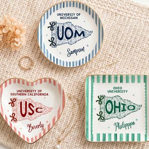 Puede incluir: Tres platos decorativos de cerámica. Uno redondo con rayas azules y texto "University of Michigan", uno en forma de corazón con texto "University of Southern California" y uno cuadrado con texto "Ohio University". Ribete dorado y diseños de banderas.