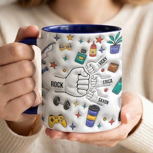 Op de afbeelding: Witte keramische mok met een blauwe binnenkant en handvat, versierd met diverse iconen en het woord "ROCK" met drie vuisten. De mok heeft ook de namen "Vicky", "Erica" en "Saxon".