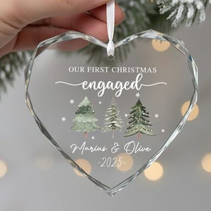 May include: Clear, heart-shaped Christmas ornament with the text "Our First Christmas engaged" and the names "Marius & Olive -2025". The ornament features three watercolor Christmas trees and is suspended by a white ribbon.